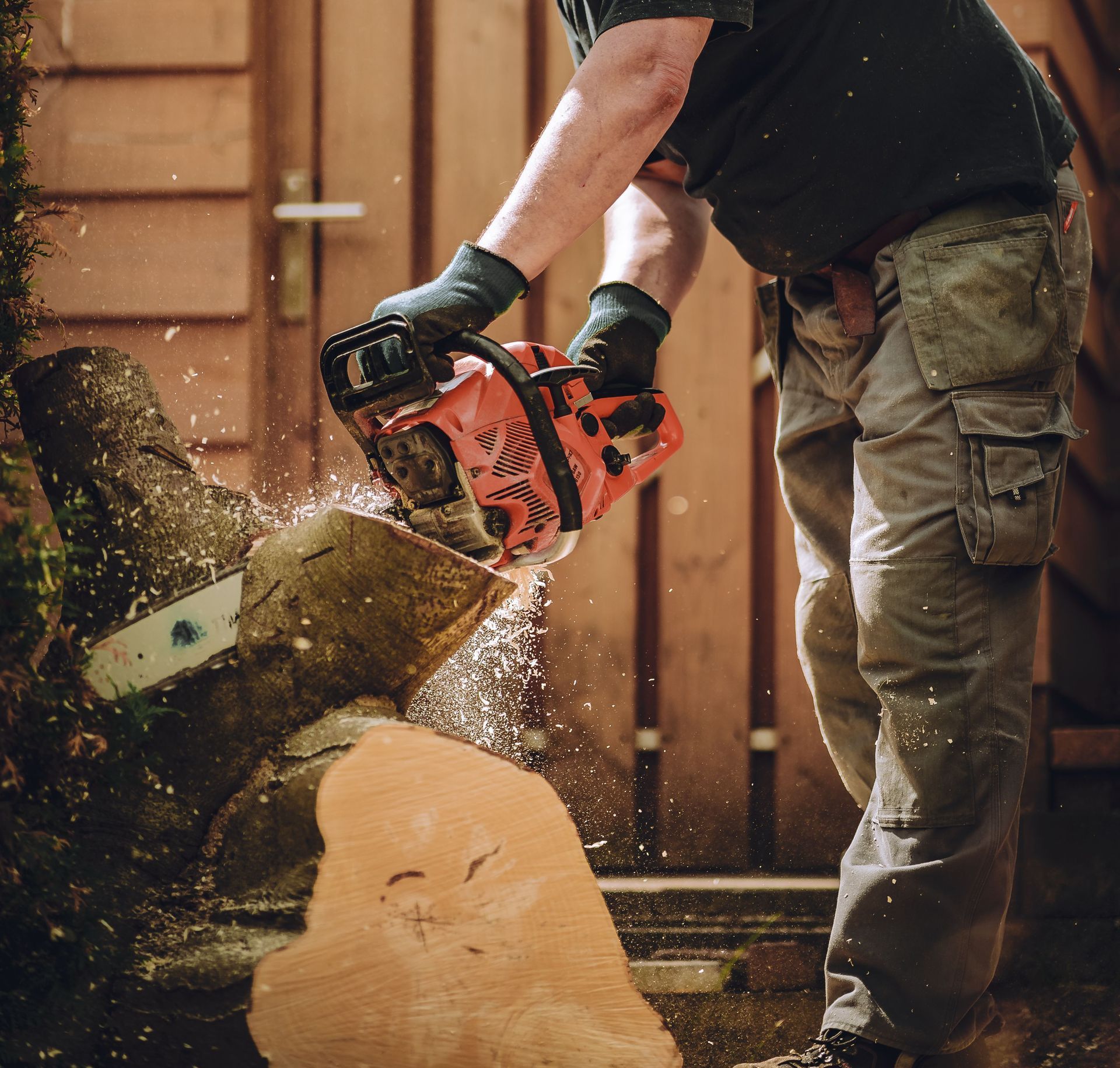 Worker using a chainsaw to cut wood, wearing safety gear in an outdoor setting.